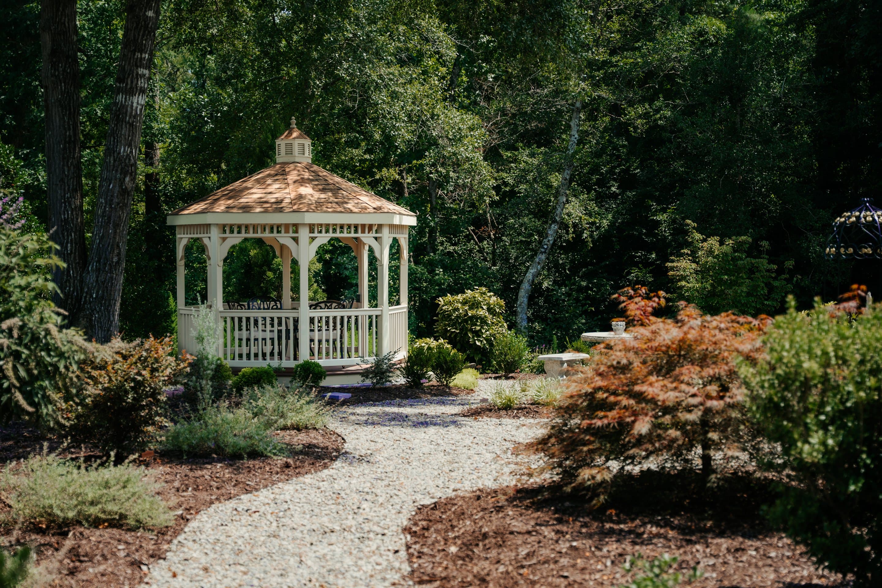 A cream gazebo in a designed garden with a curved gravel path.