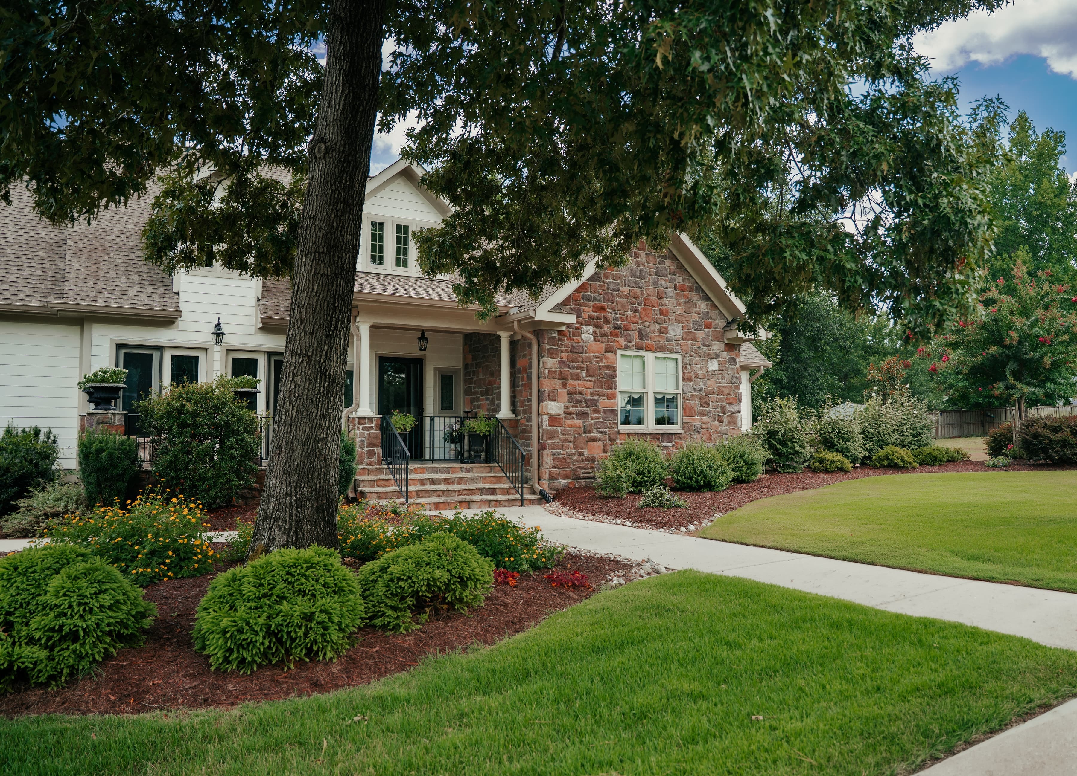 A stone-and-siding home with a manicured lawn, curved planting beds, and a shaded oak tree.