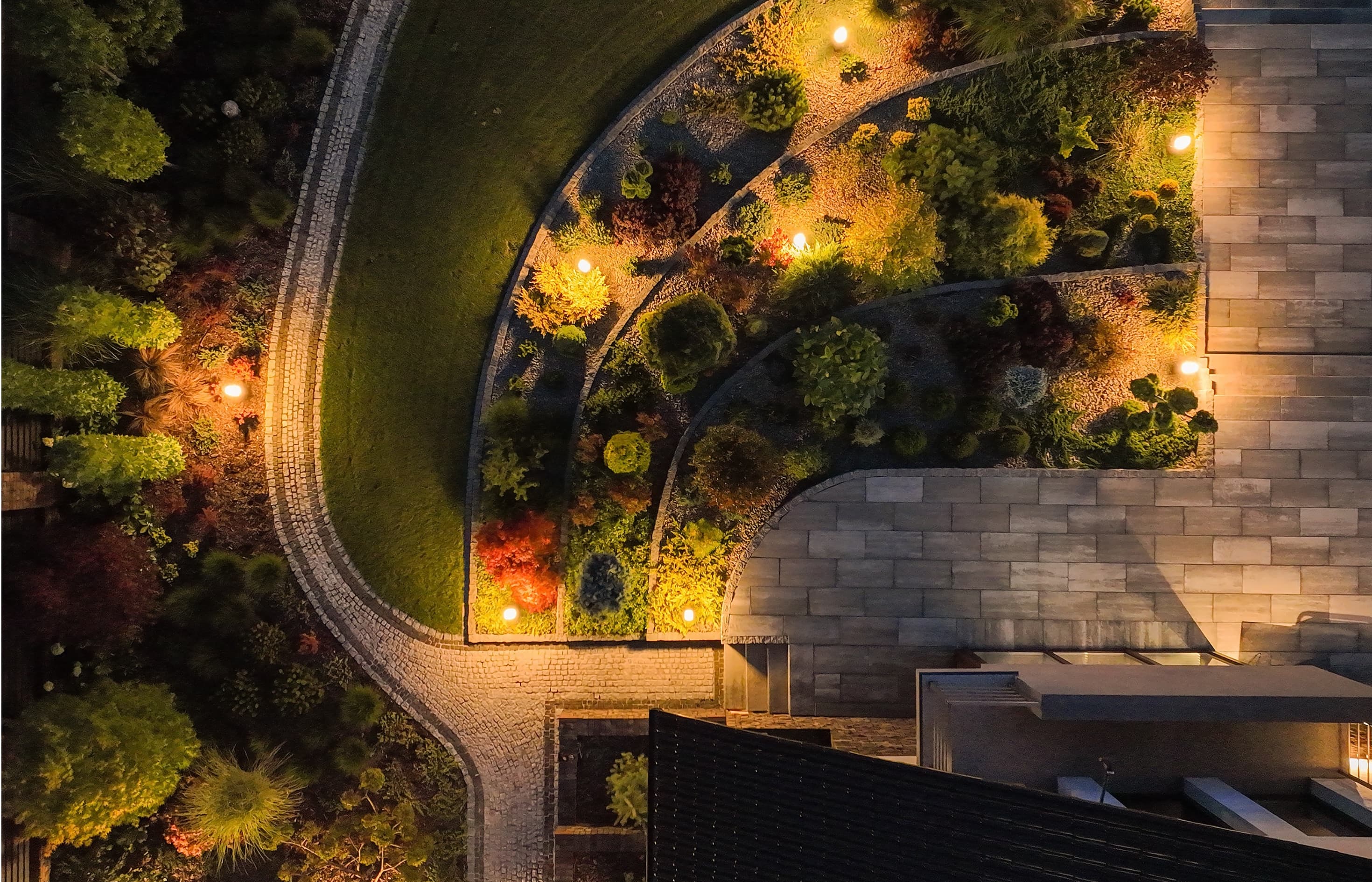 An aerial dusk view of curved paver walls, cobblestone paths, and lit planting beds around a modern home.
