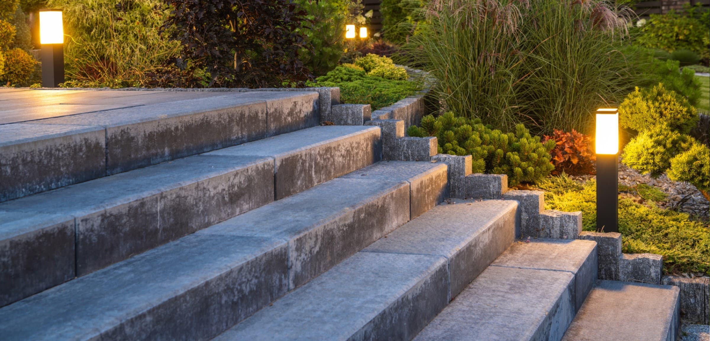 Paver stone steps at dusk lit by low bollard lights beside layered planting beds.