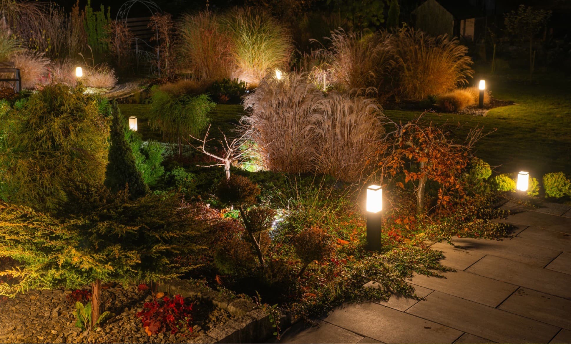 A garden bed at night lit by warm low-voltage bollard lights along a paver path.